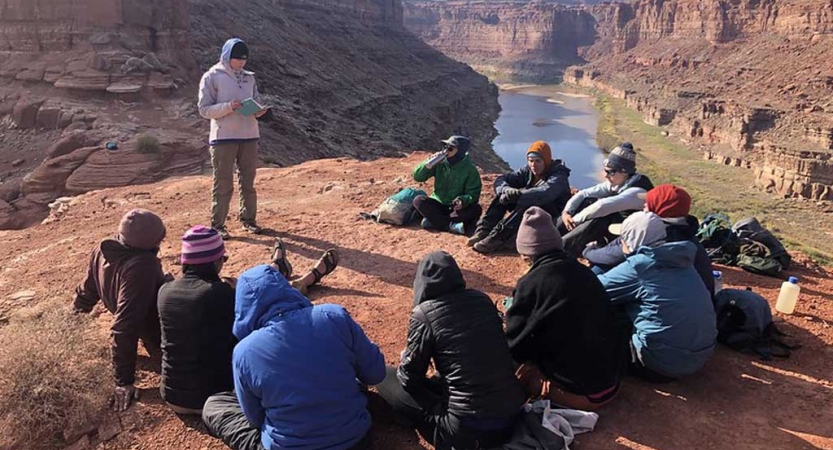 a group of students sit atop a rock high above a river listening to a reading from the outward bound readings book